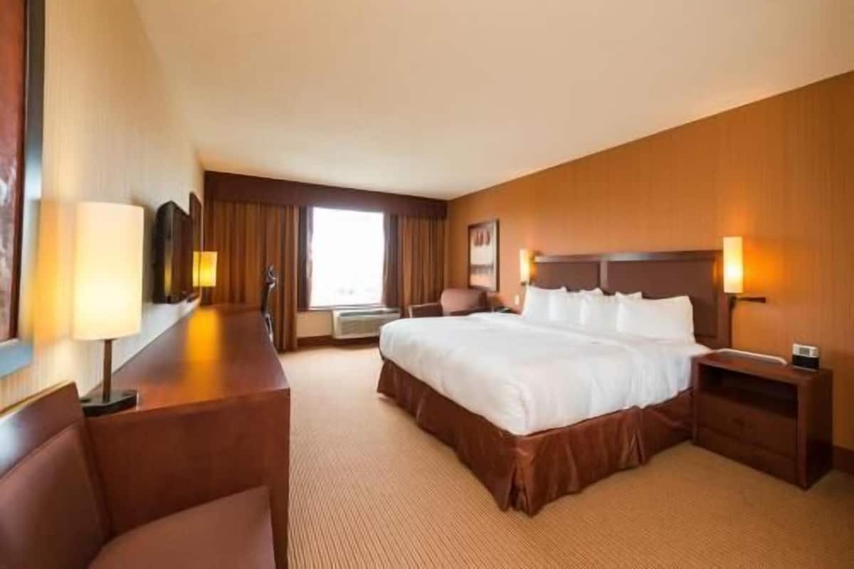 A hotel room with a large bed, a desk with a chair, a bedside table, and a window with curtains.