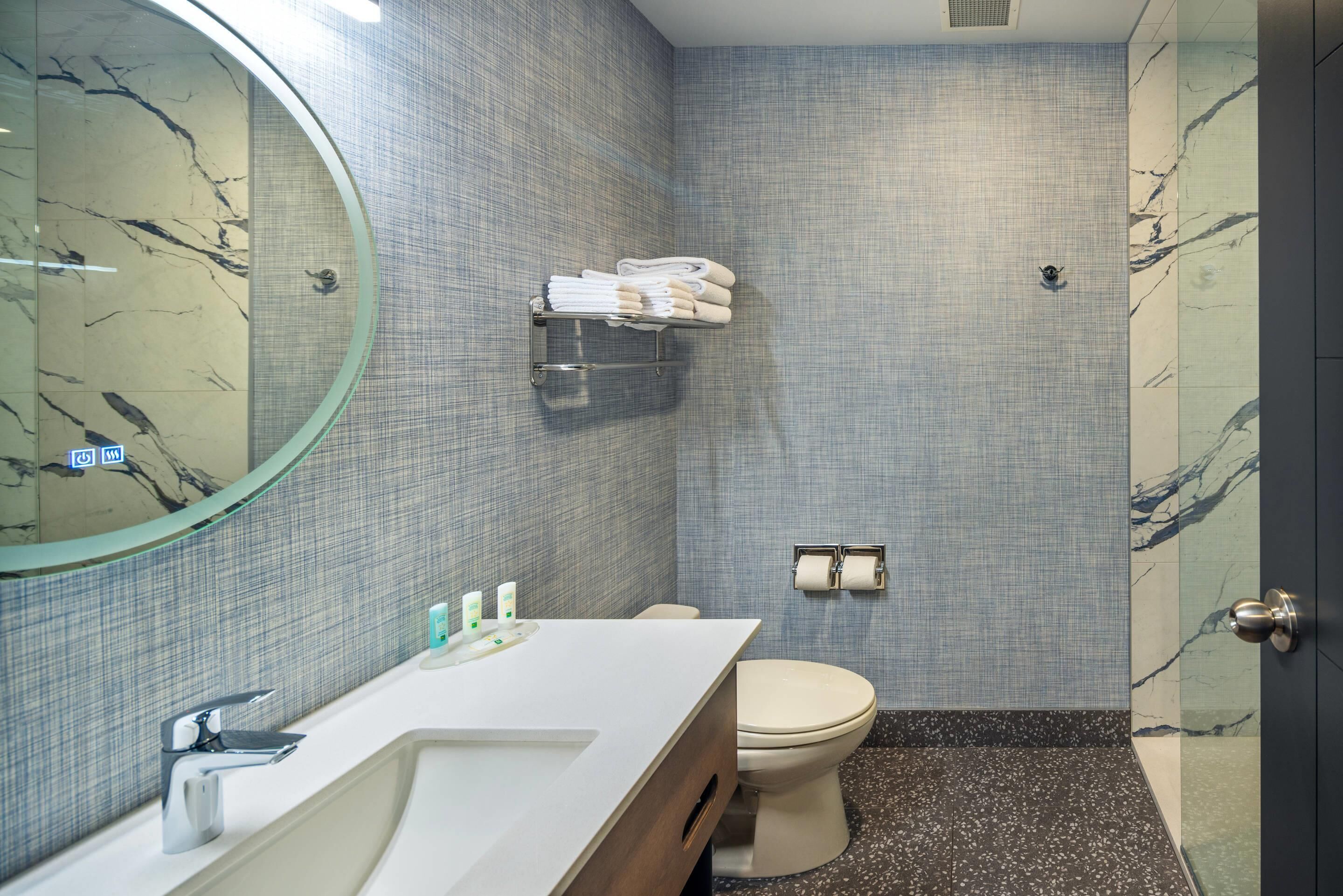 Un baño con pared de azulejos de mármol, un lavamanos blanco con espejo, un inodoro y un perchero para toallas.