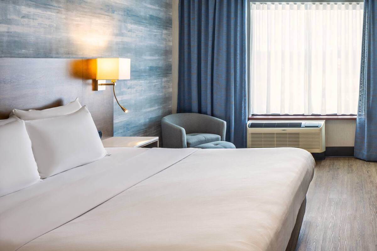 A hotel room with a large bed, a nightstand, a lamp, a chair, and a window with curtains.