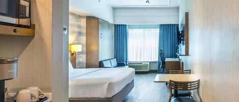 A hotel room with a bed, a desk, a chair, a microwave, and a coffee machine.