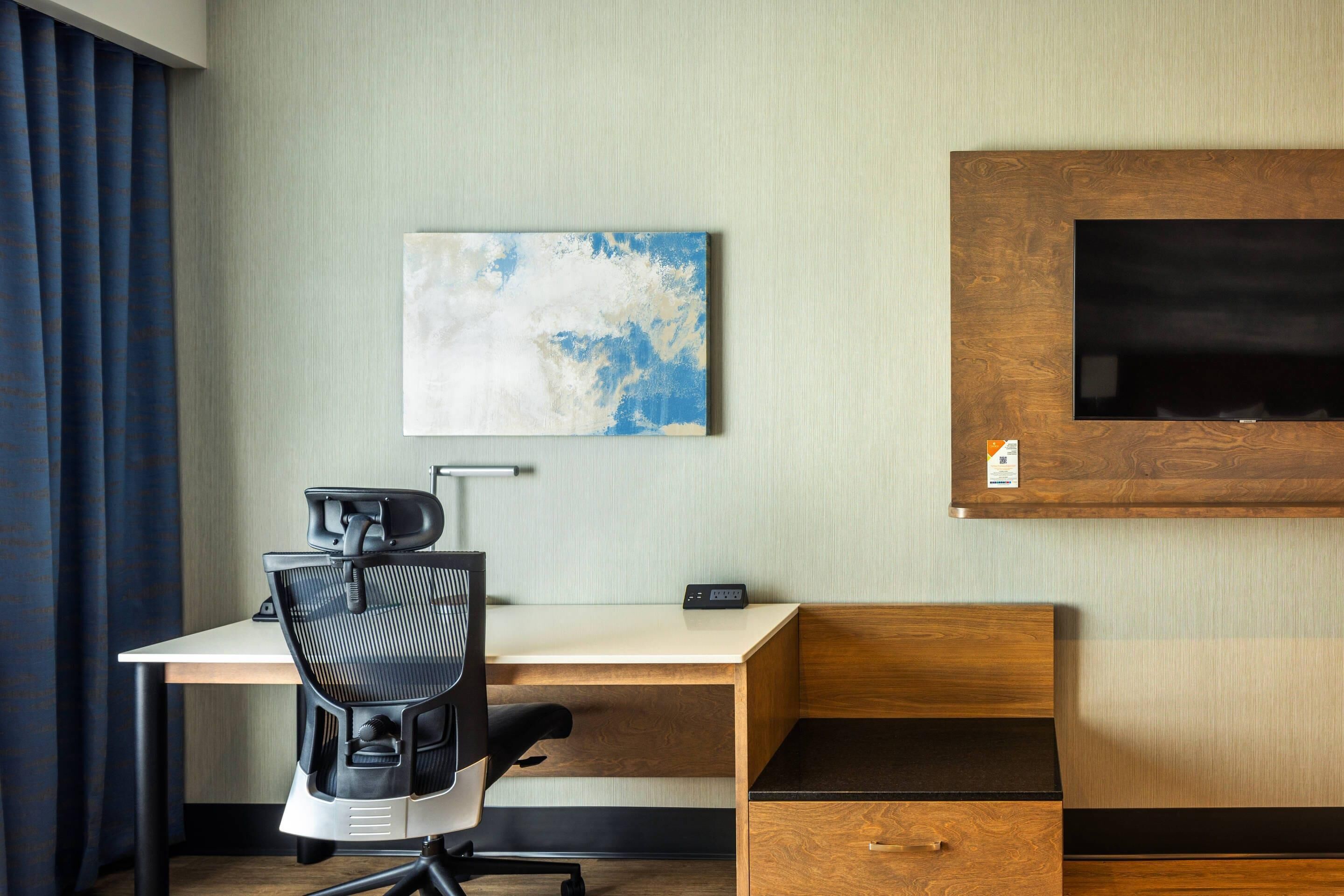 A hotel room with a wooden desk, a black office chair, a flat-screen TV mounted on a wooden panel, and a wall-mounted artwork.