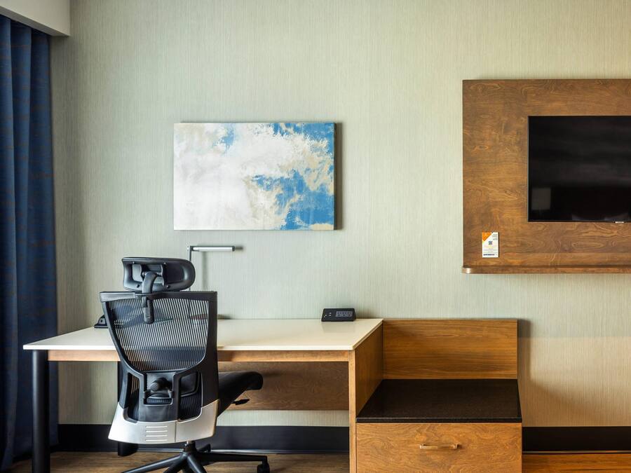 A hotel room with a wooden desk, a black office chair, a flat-screen TV mounted on a wooden panel, and a wall-mounted artwork.