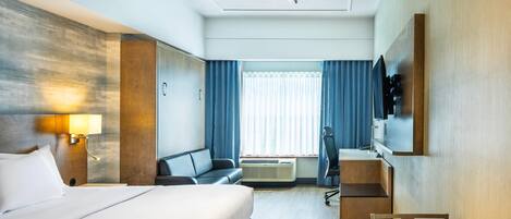 A hotel room with a bed, a desk, a chair, a sofa, a TV, and a window with curtains.