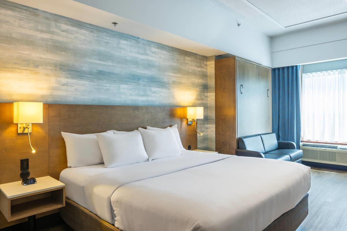 A hotel room with a large bed, two bedside lamps, a nightstand, a seating area with a chair, and a large window with curtains.