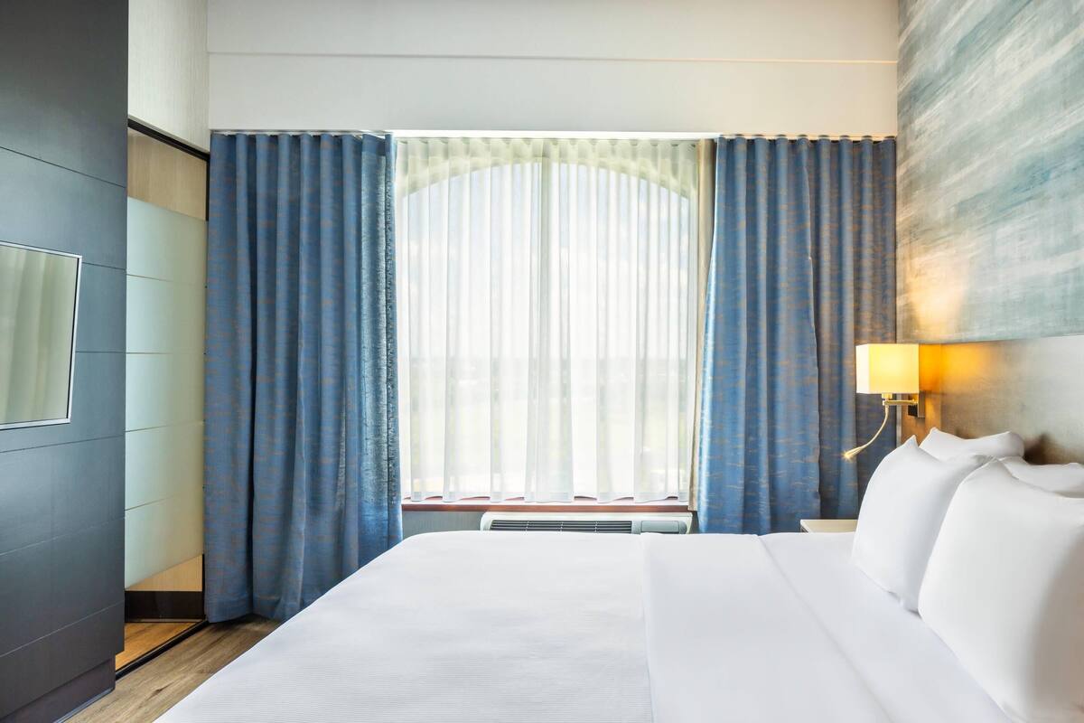 A hotel room with a large bed, a window with curtains, a wall-mounted lamp, and a radiator.