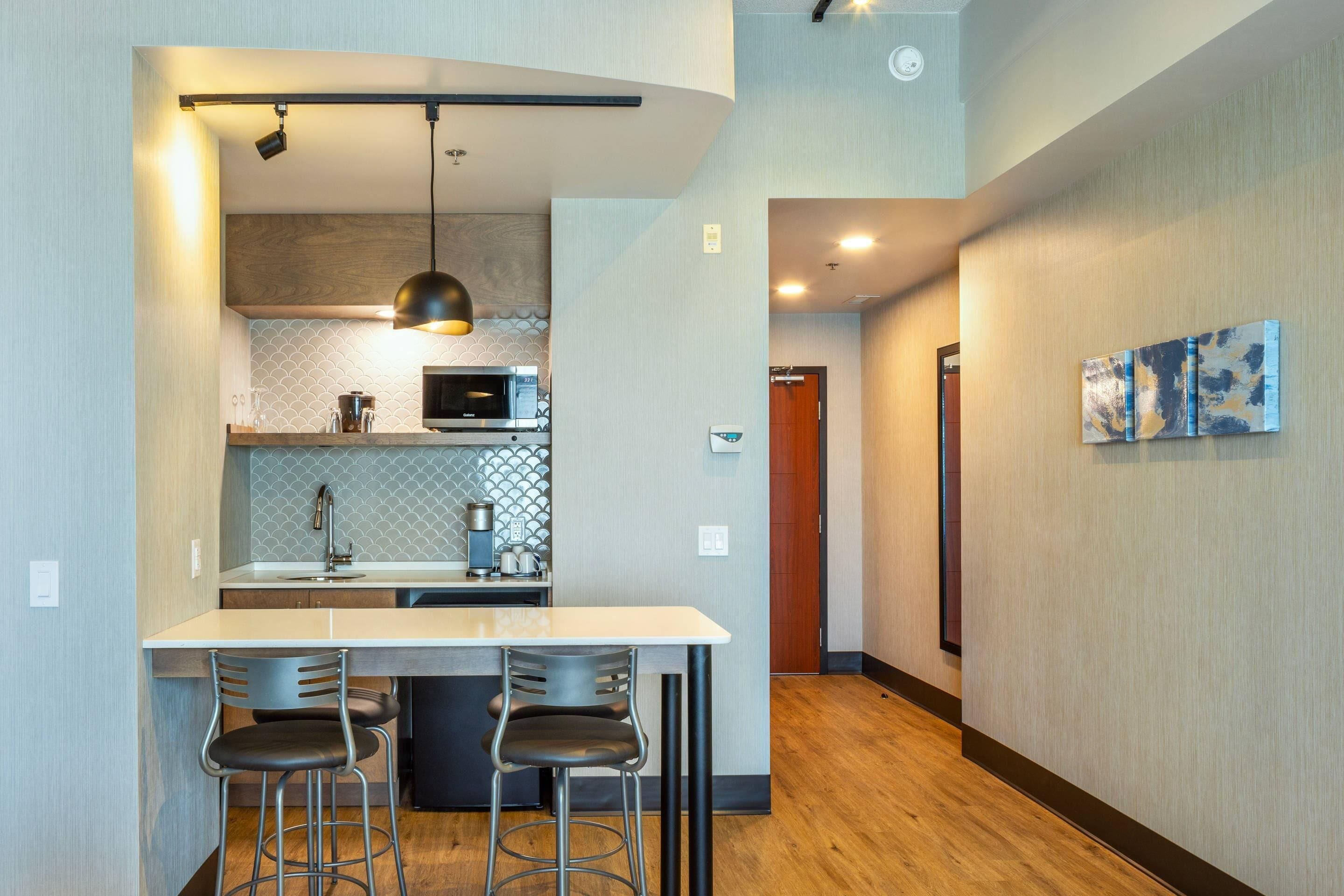 A modern kitchen with a white countertop, two bar stools, a pendant light, and a microwave.