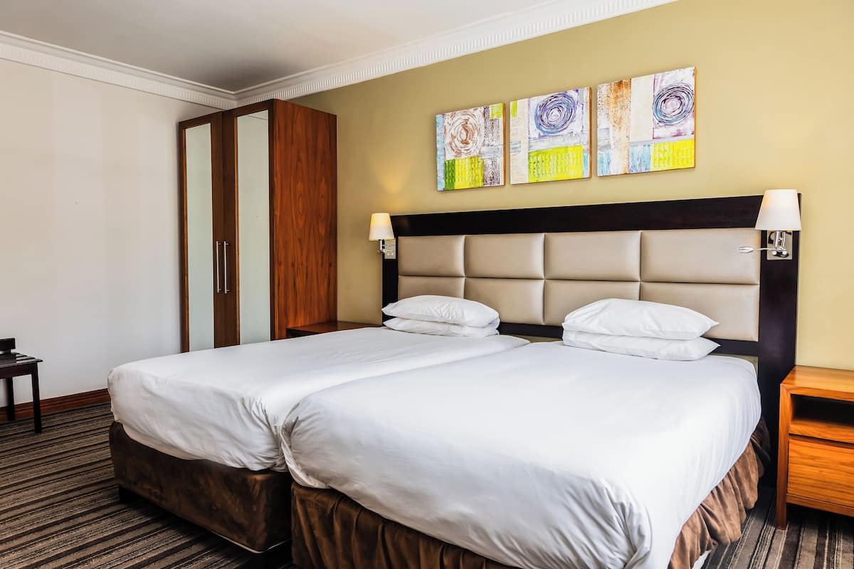 A hotel room with two beds, a wooden wardrobe, and abstract wall art.