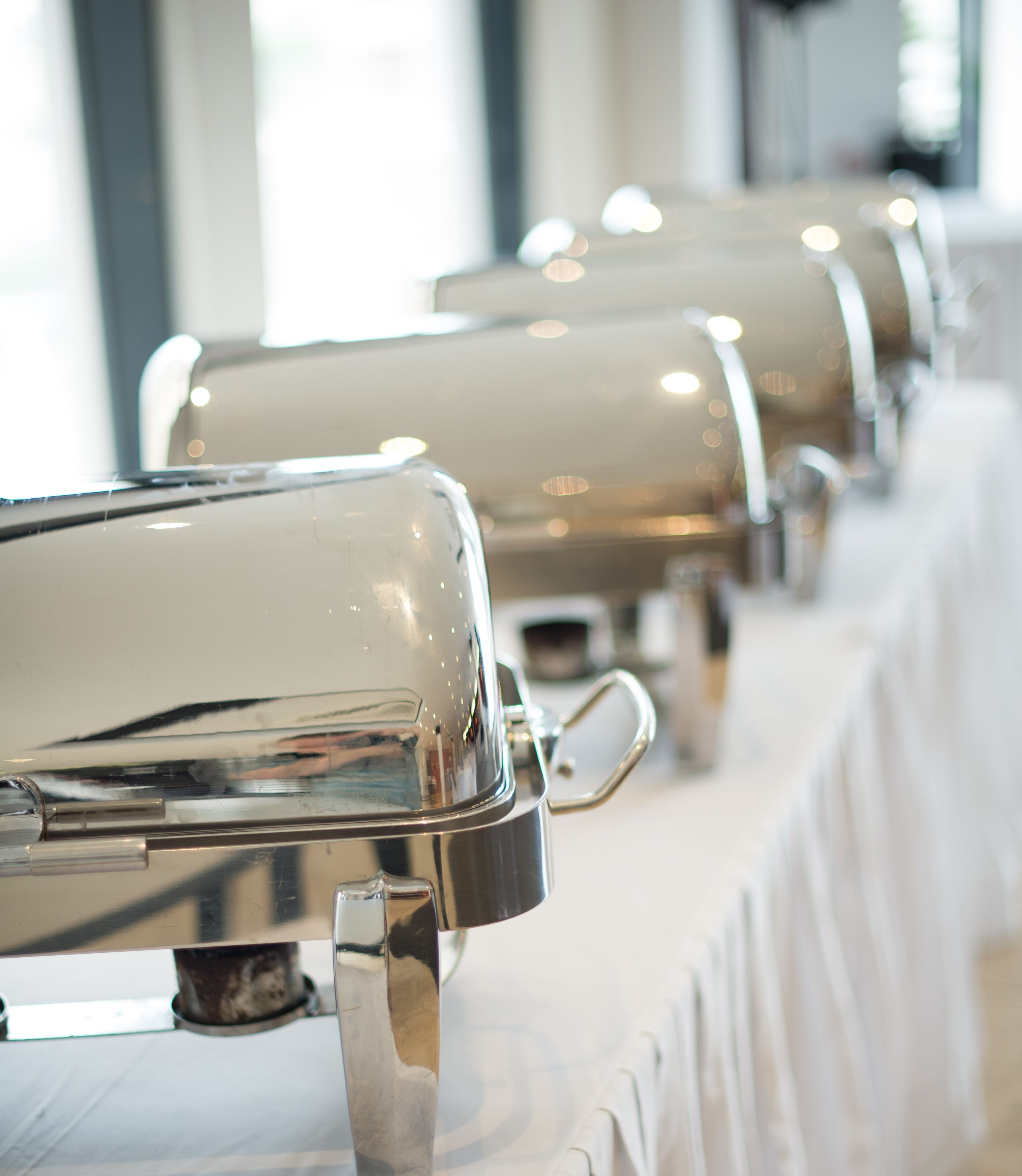 A row of buffet service stations with metallic tops and handles, set against a white tablecloth.