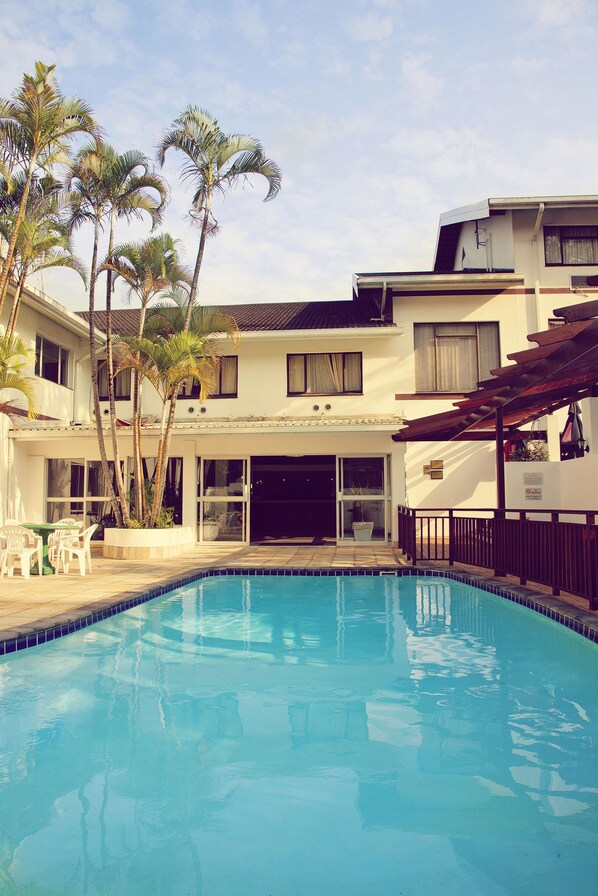 A swimming pool with a clear blue water, surrounded by palm trees and a building with a covered patio area.