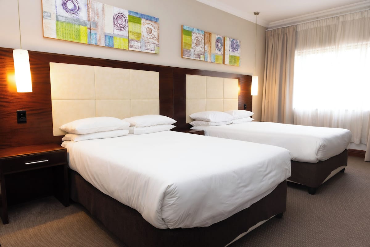 A hotel room with two beds, a wooden headboard, a nightstand, and artwork on the wall.