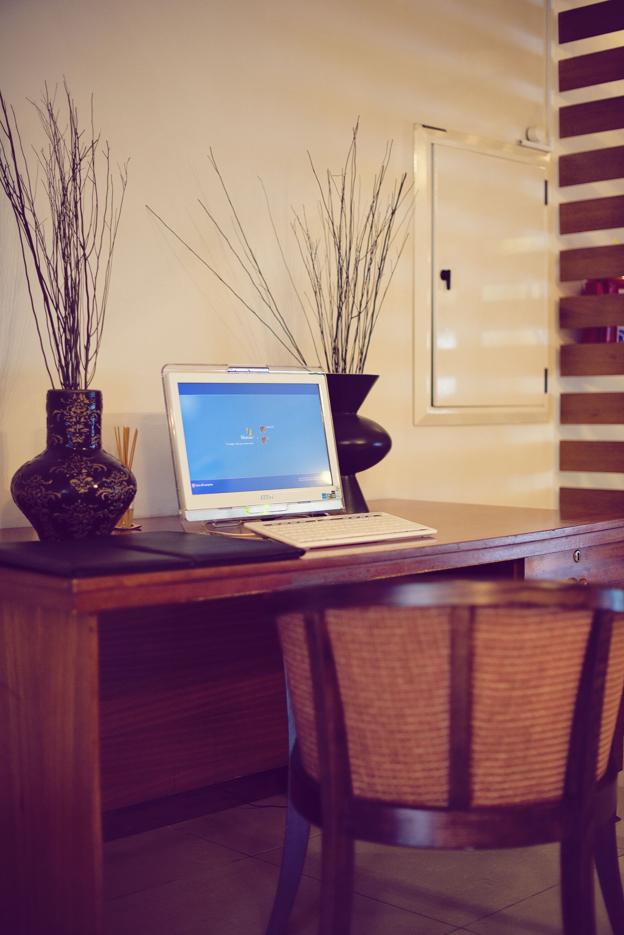 A wooden desk with a laptop, two vases with branches, and a wicker chair.