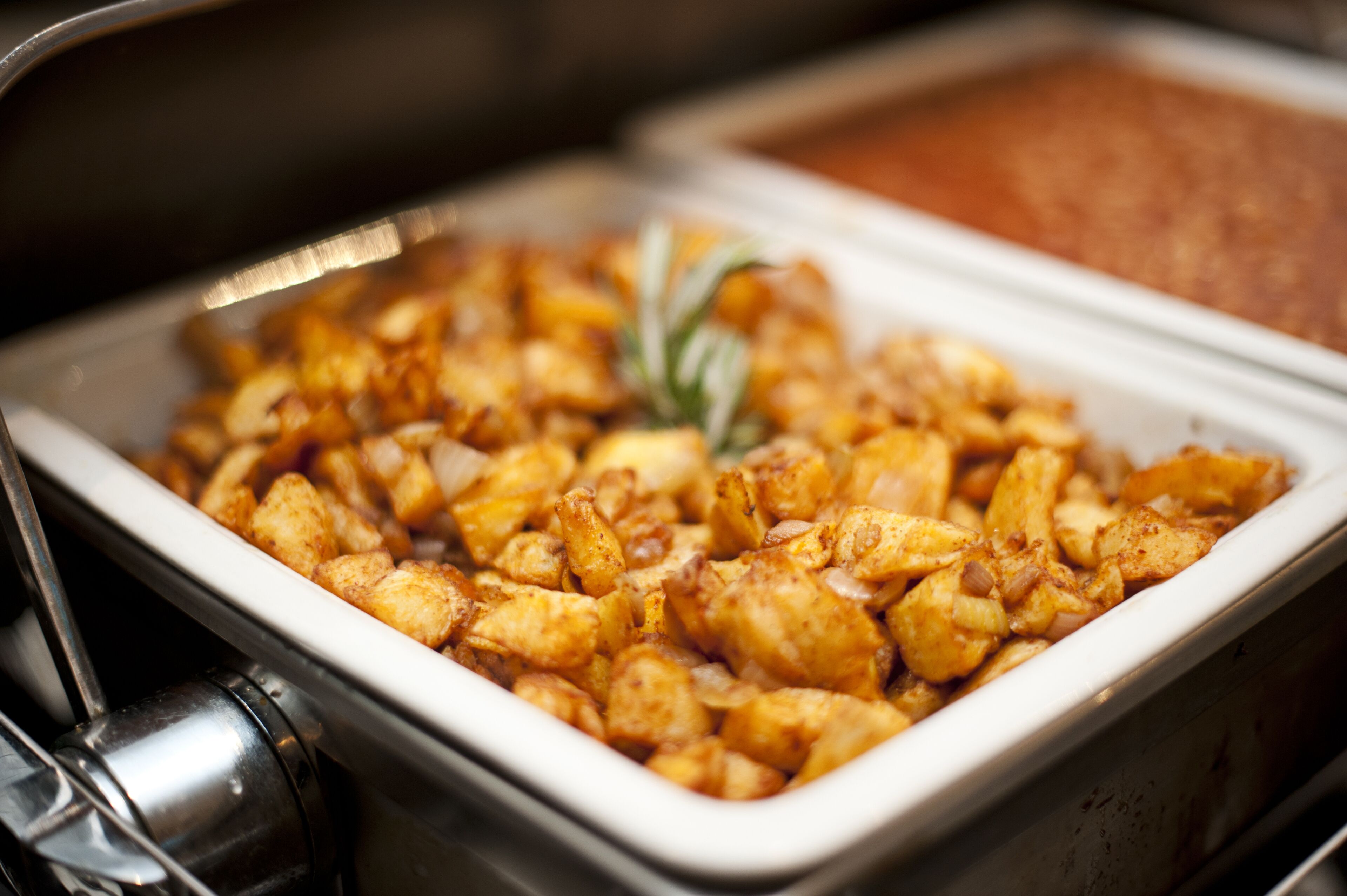 A buffet with a variety of dishes, including a tray of seasoned potatoes and a container of sauce.