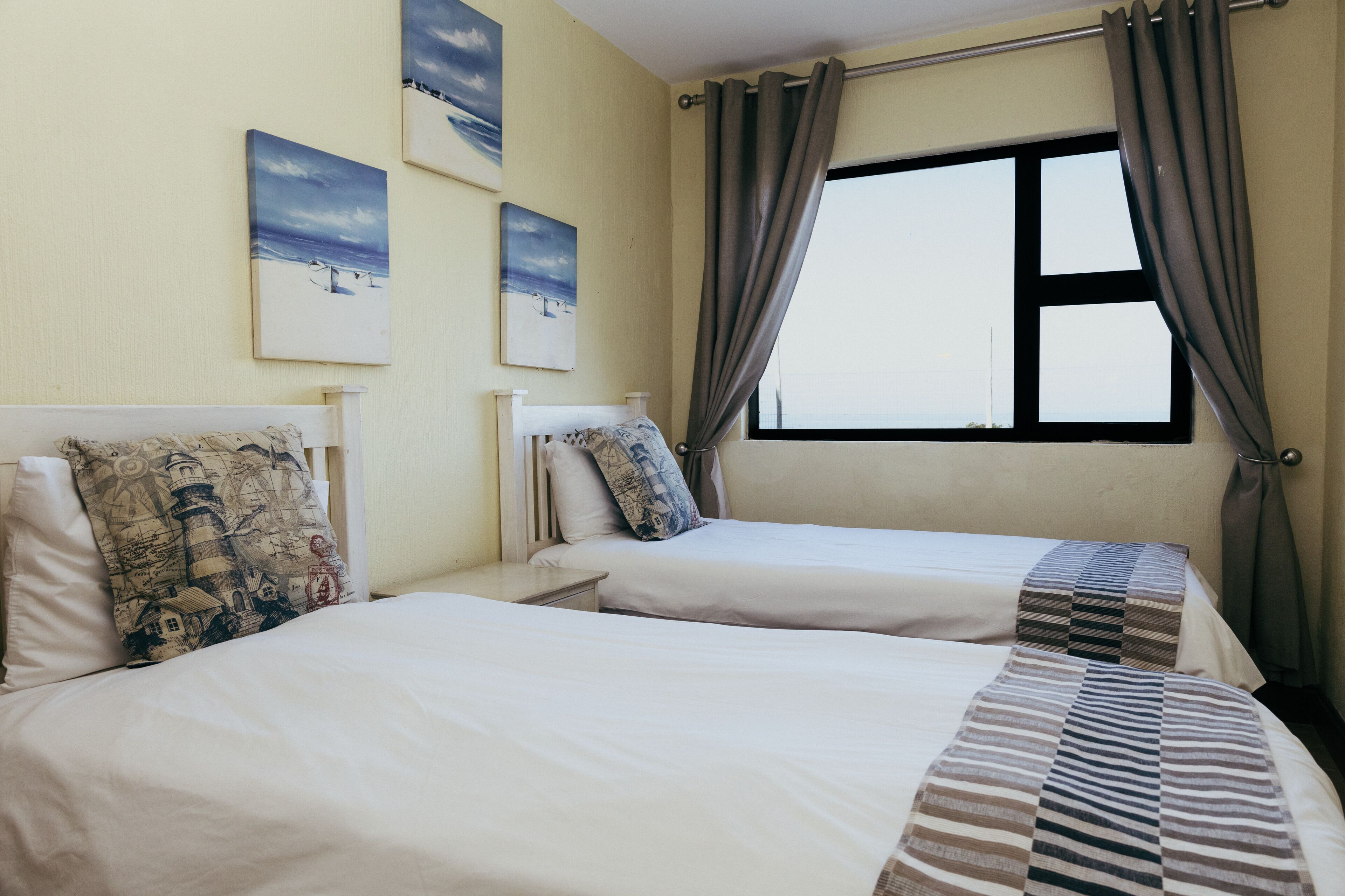 A hotel room with two beds, a large window with curtains, and three paintings on the wall.