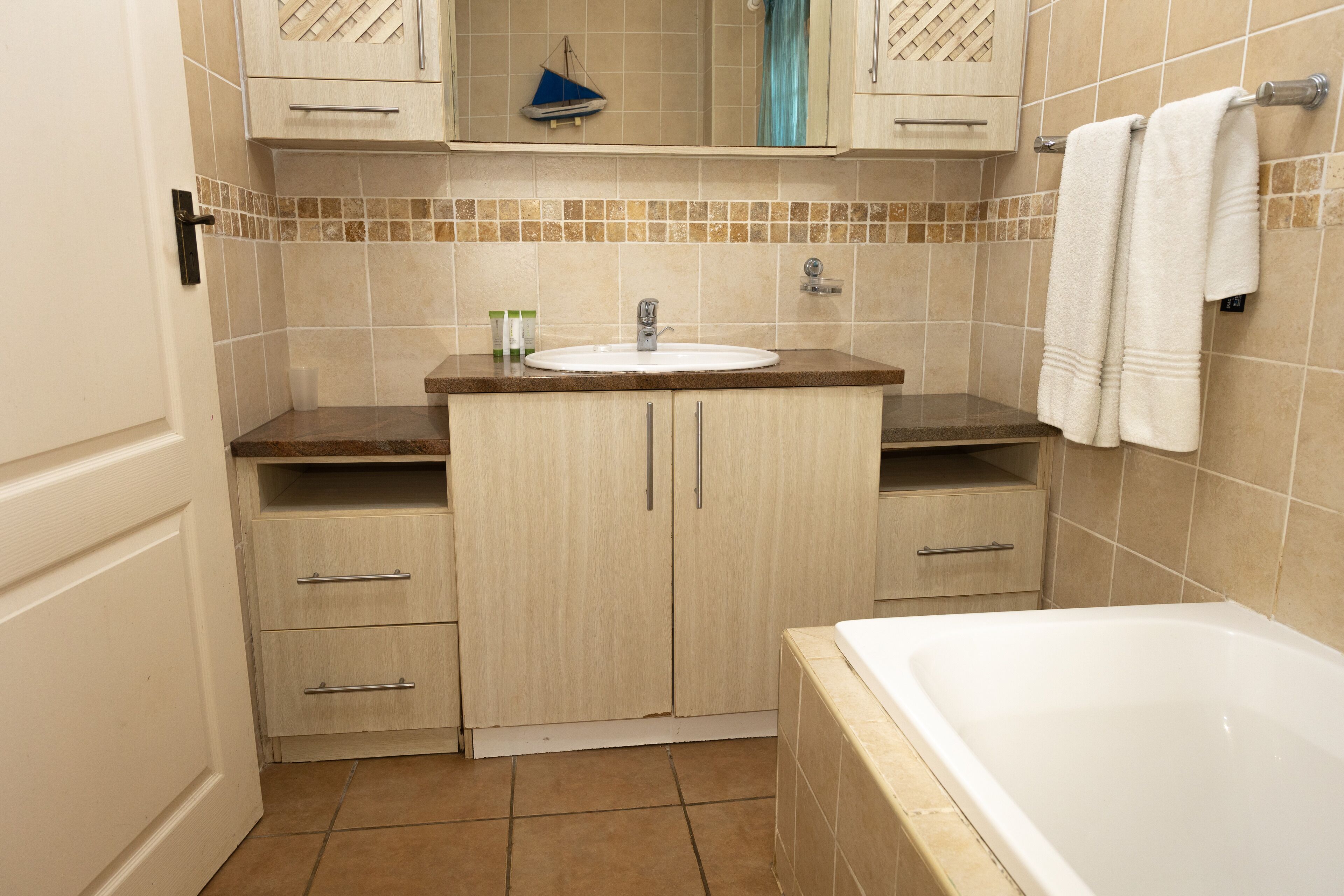 Un baño con lavabo, espejo y bañera. Hay armarios y un perchero para toallas.