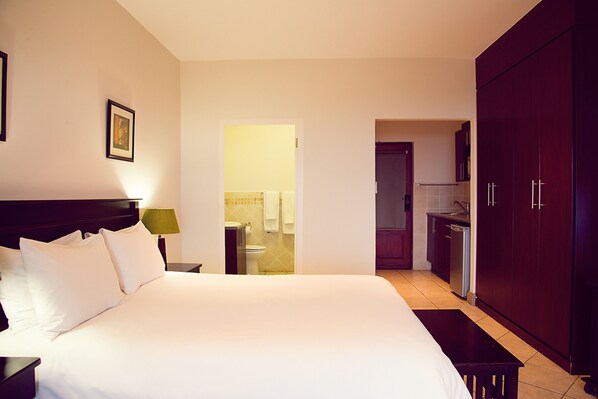 A hotel room with a large bed, a nightstand, a desk, a wardrobe, and a view into the bathroom and kitchen. - The Edwardian (Port Edward)