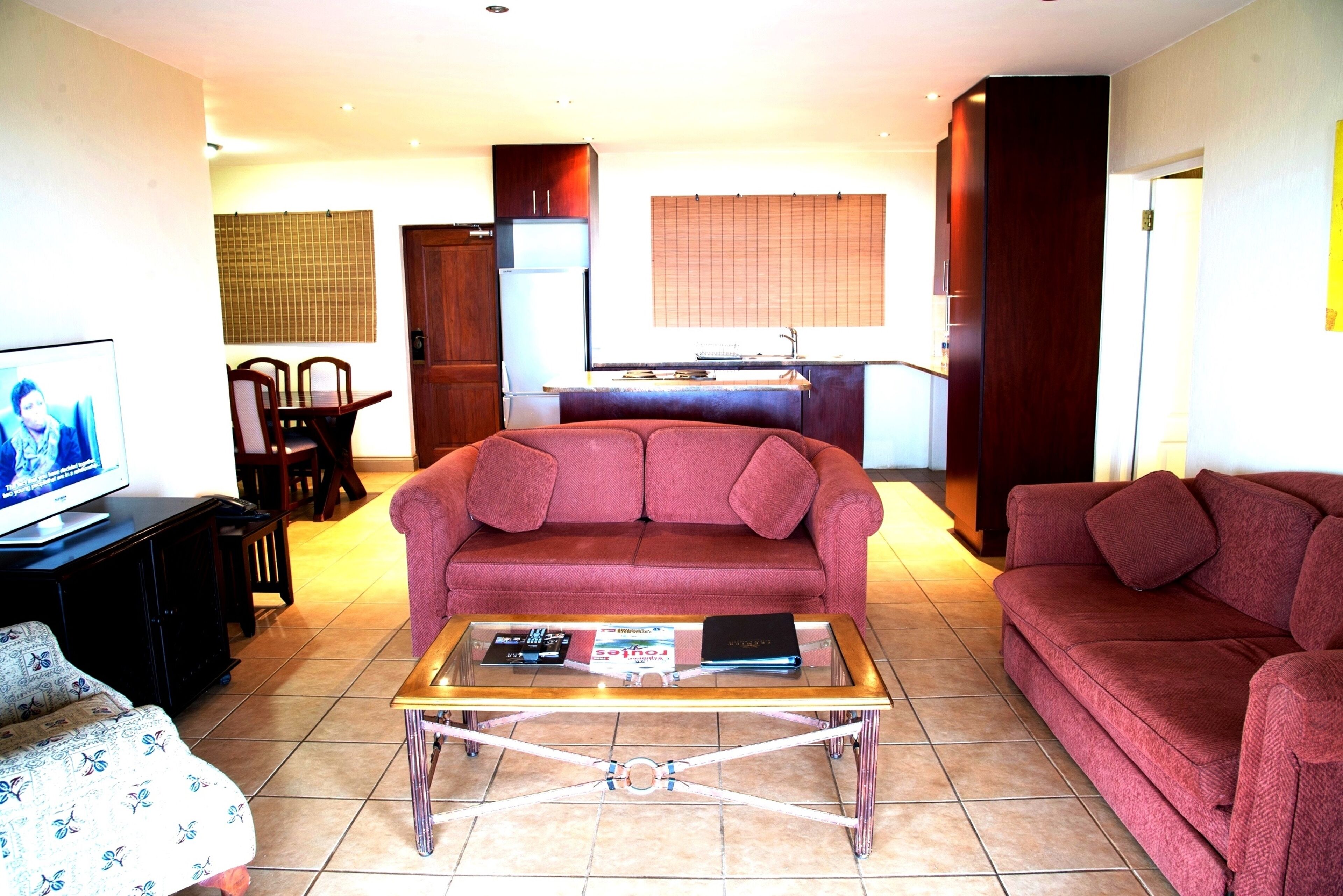 A living room with a sofa, a coffee table, and a TV. There is a kitchen in the background with a refrigerator and a window.
