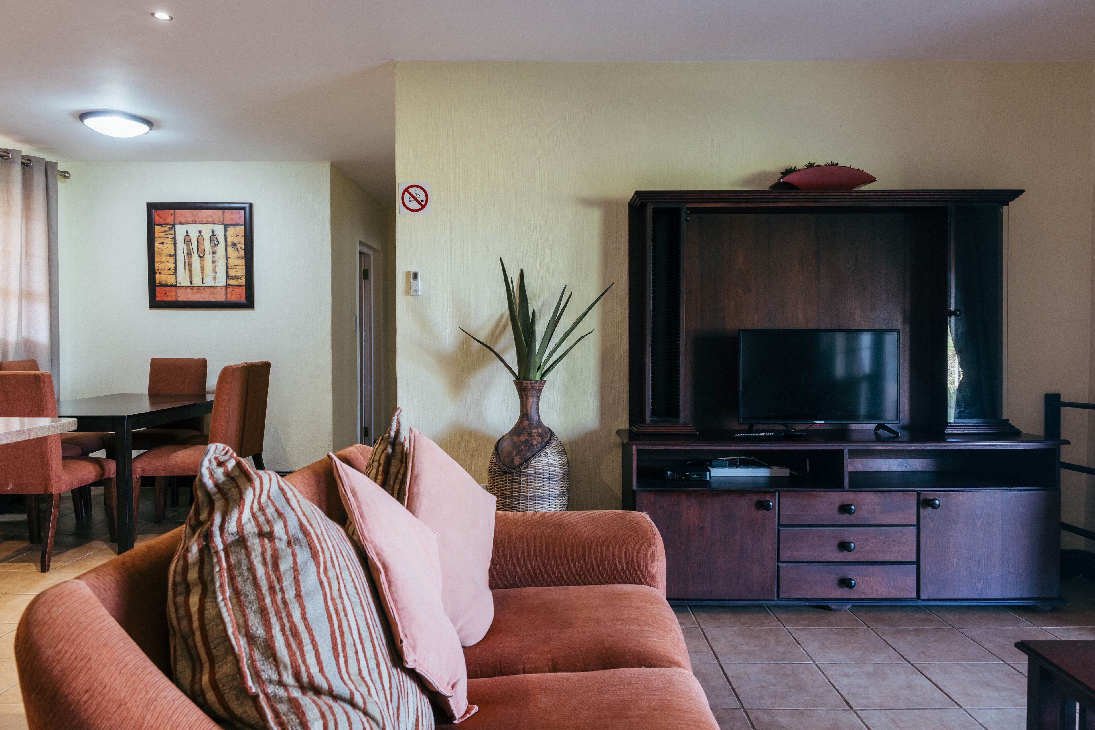 A living room with a sofa, a TV on a wooden cabinet, a dining area with chairs, and a vase with plants.