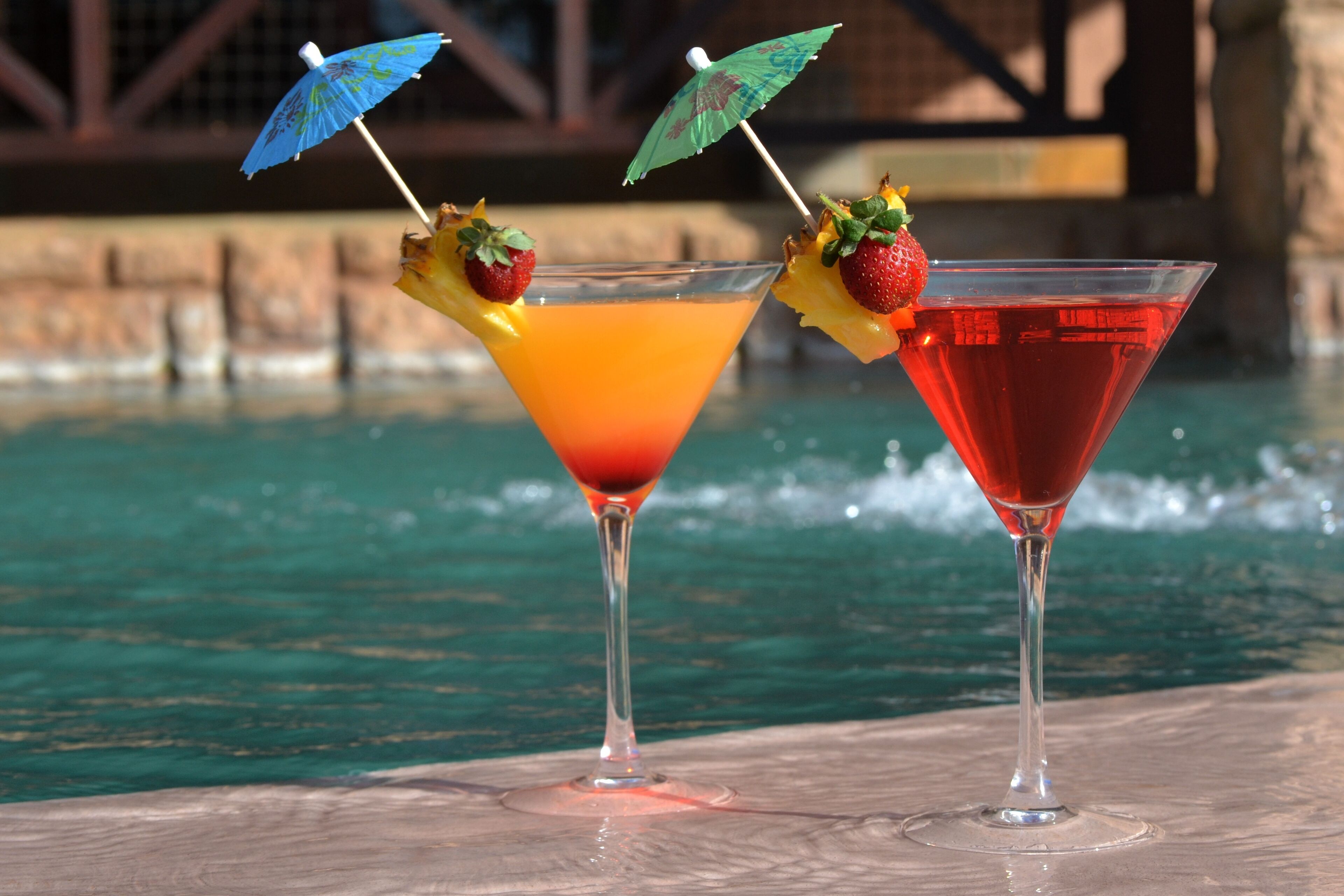 Two cocktails with umbrellas and fruit garnishes by a poolside.