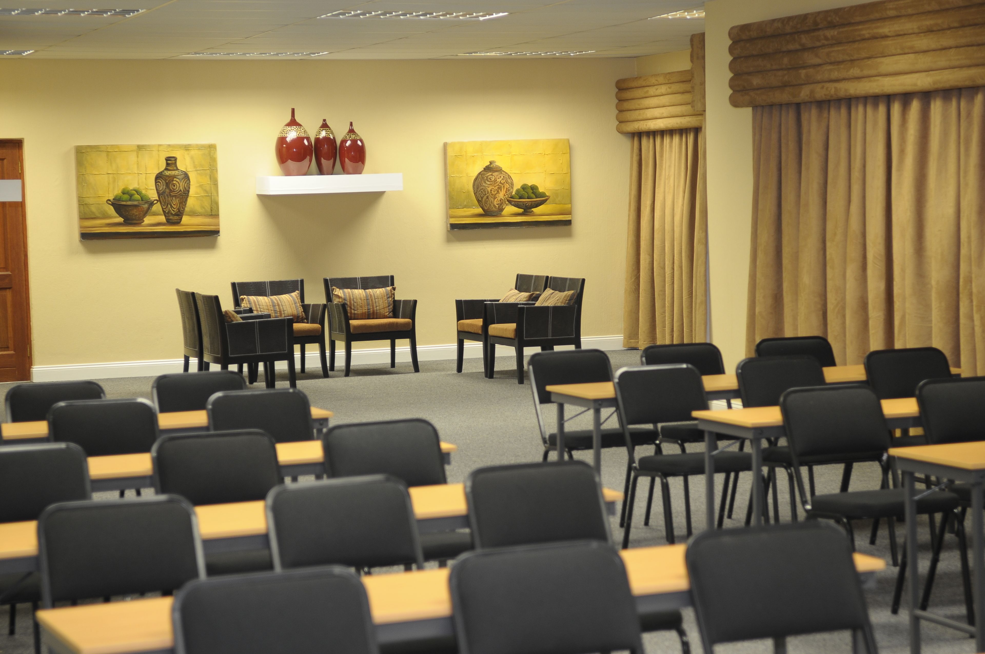 Un salón de conferencias con filas de sillas y mesas, dos cuadros en la pared y una cortina color beige.