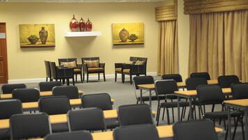 Un salón de conferencias con filas de sillas y mesas, dos cuadros en la pared y una cortina color beige.