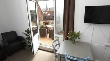 A room with a sliding glass door leading to an outdoor balcony with a table and chairs, a TV mounted on the wall, and a small table with a vase of flowers.