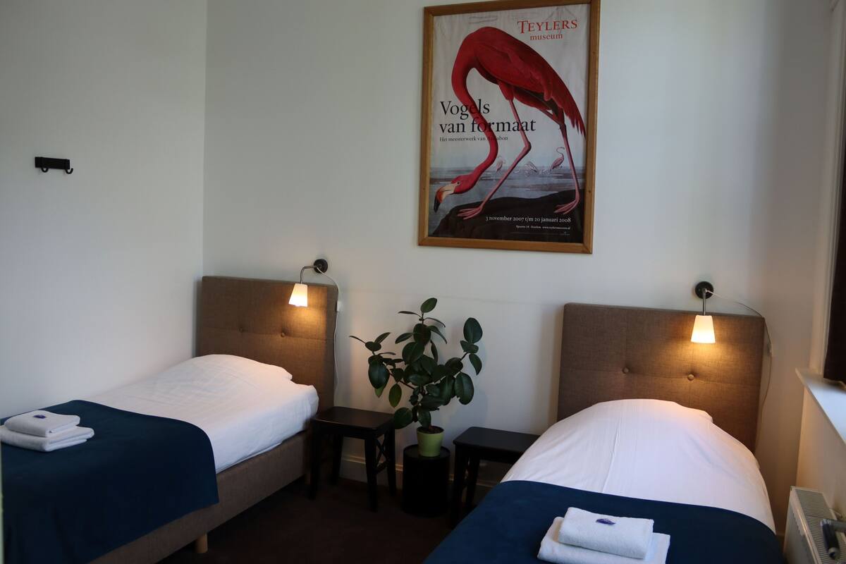 A hotel room with two beds, a nightstand, a potted plant, and a poster on the wall.