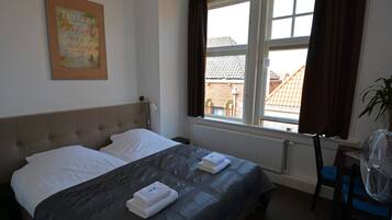 A neatly made bed with white linens and a dark bedspread, a framed picture on the wall, a window with curtains, and a radiator.