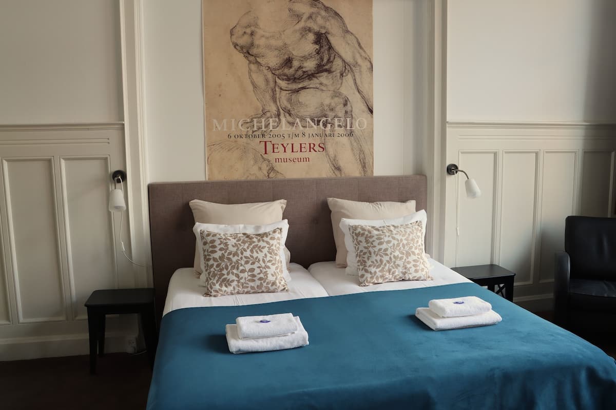 A hotel room with a bed, pillows, a blue bedspread, a wall-mounted artwork, and a nightstand.