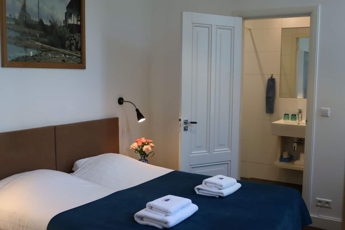 A hotel room with a bed, a nightstand with a lamp, a vase of flowers, and a bathroom visible through an open door.
