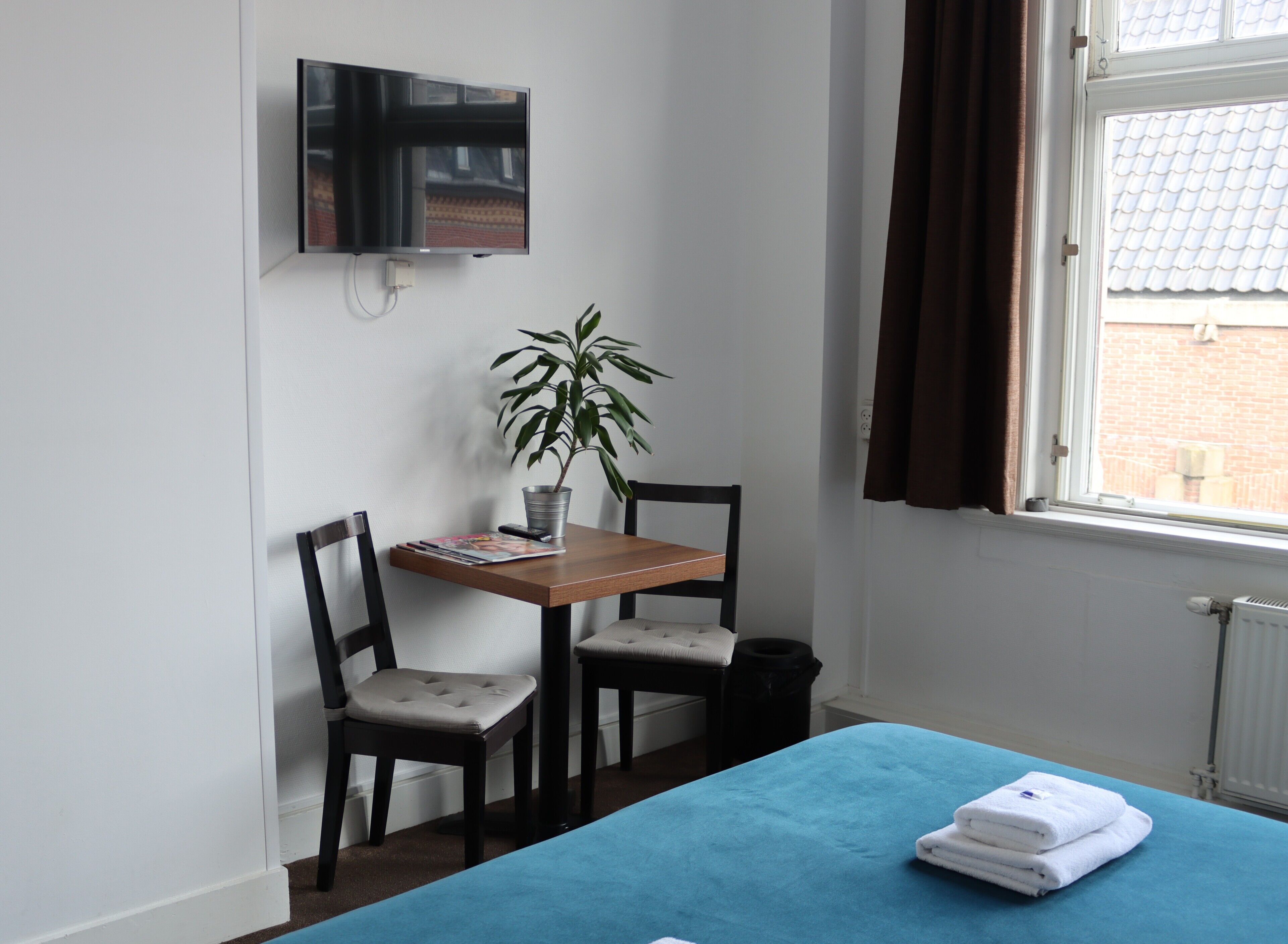 Una habitación pequeña con cama, un escritorio con silla, televisor montado en la pared y una ventana con cortinas.