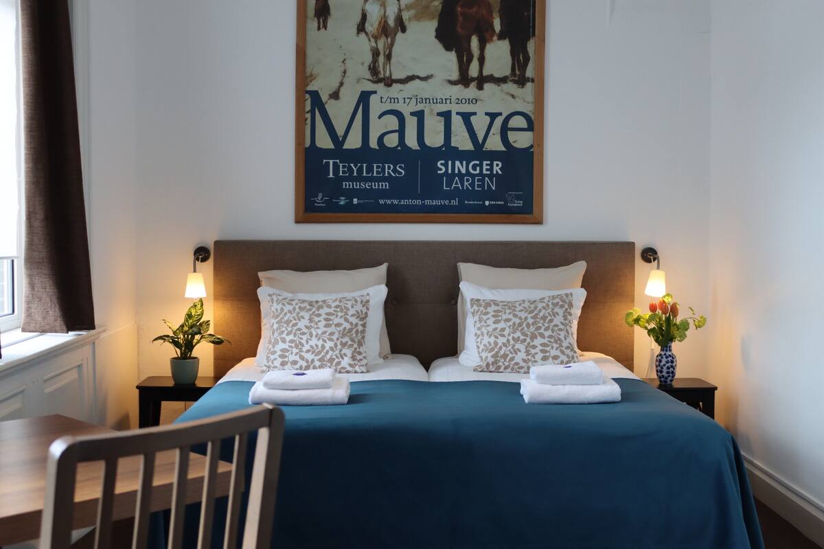 A hotel room with a bed, two nightstands, a potted plant, and a poster on the wall.