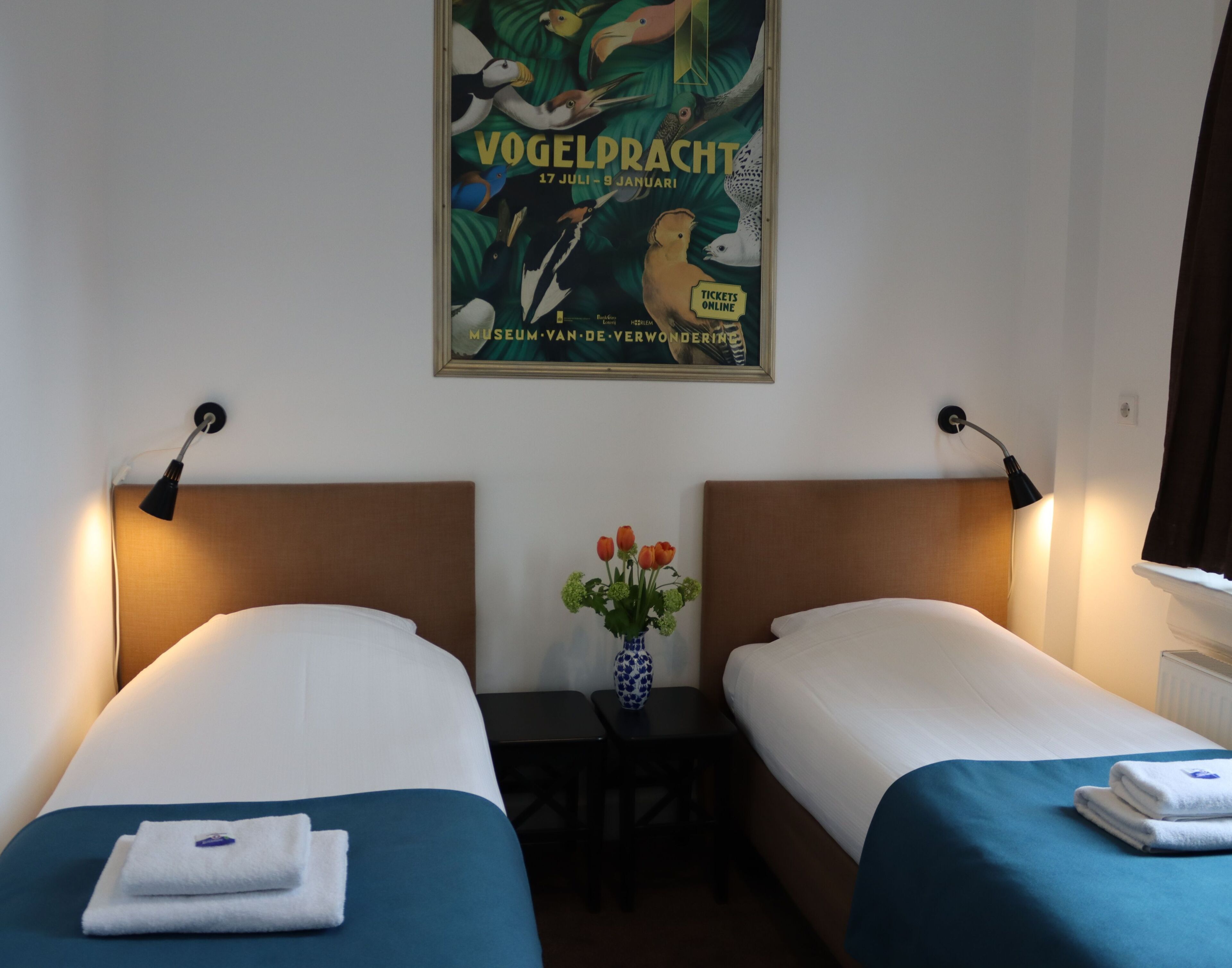 A hotel room with two beds, a small table with a vase of flowers, and a poster on the wall.