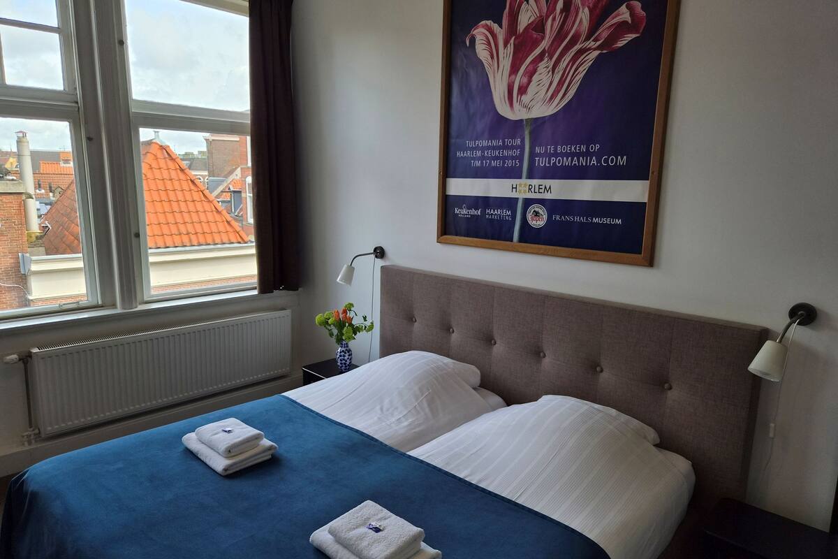 A hotel room with a bed, a window with curtains, a wall-mounted poster, and a bedside table with a lamp.
