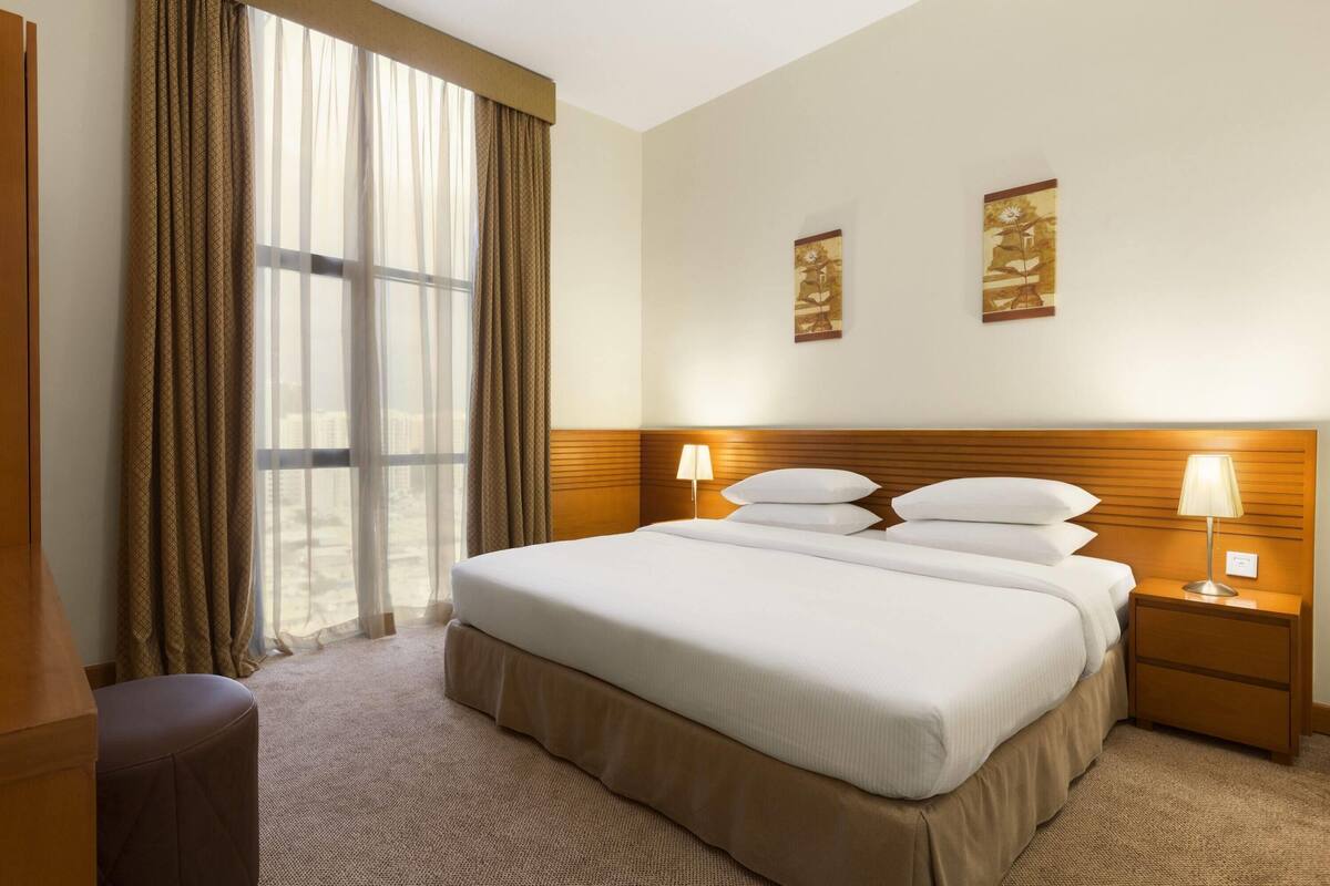 Une chambre d’hôtel avec un grand lit, deux lampes de chevet, une tête de lit en bois et une fenêtre avec des rideaux.