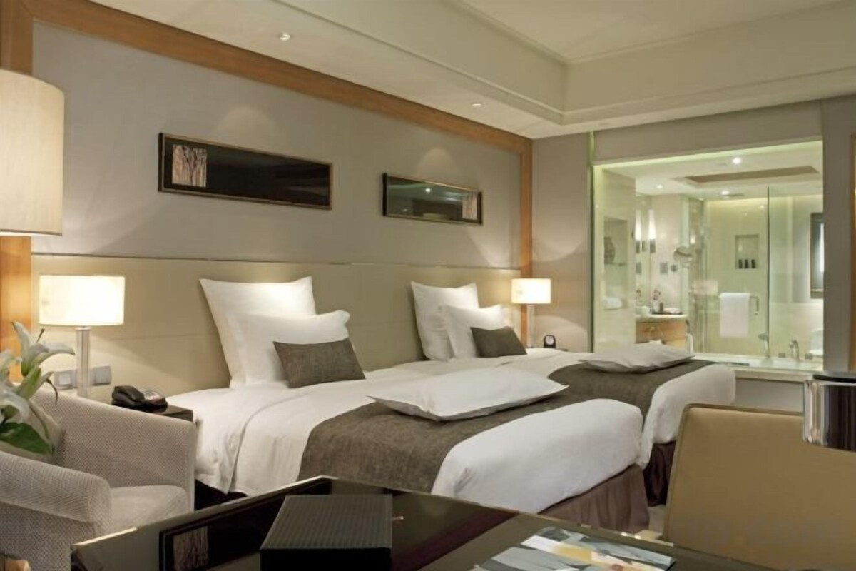 A hotel room with two beds, a desk, a chair, a mirror, and a bathroom visible through the glass door.