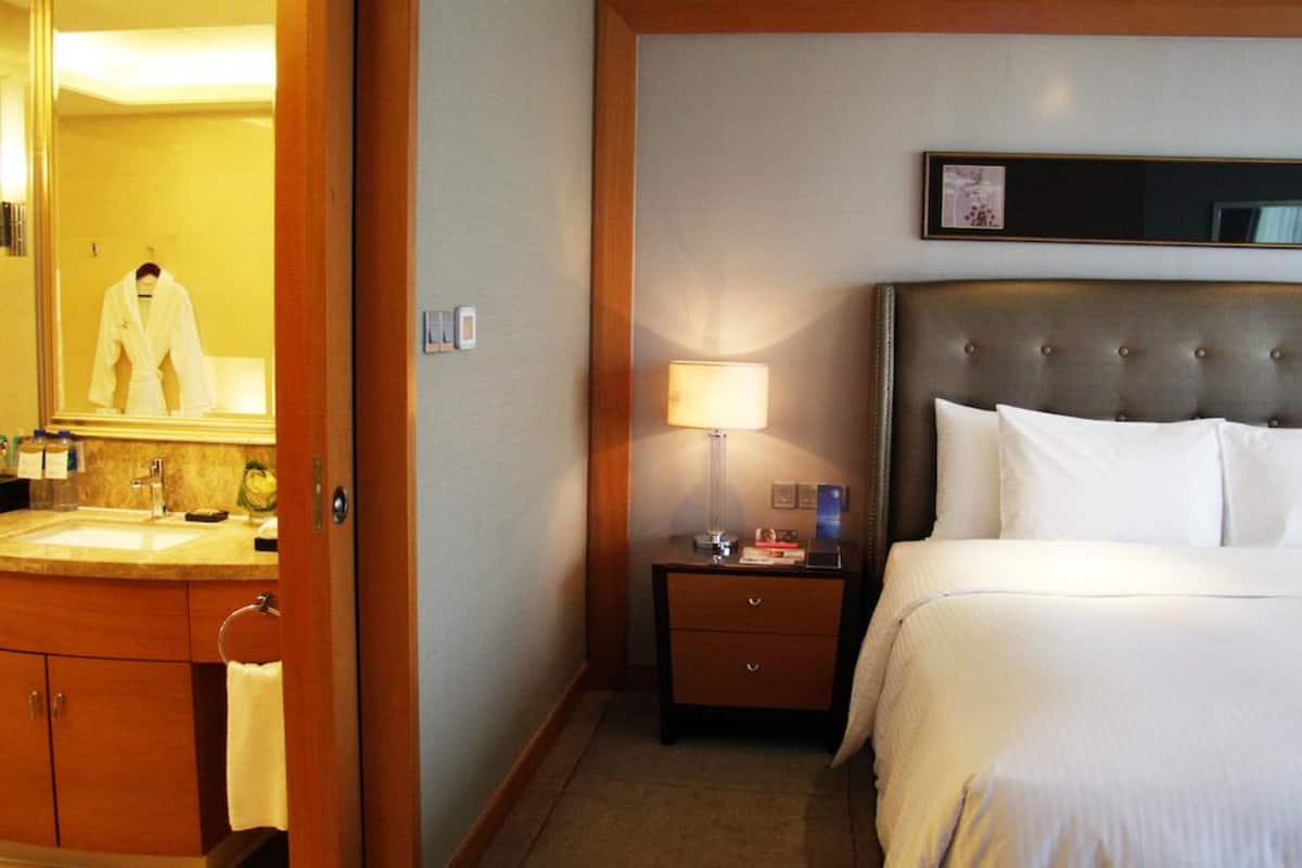A hotel room with a large bed, a bedside table with a lamp, a nightstand, a bathroom with a sink and mirror, and a robe hanging on the door.
