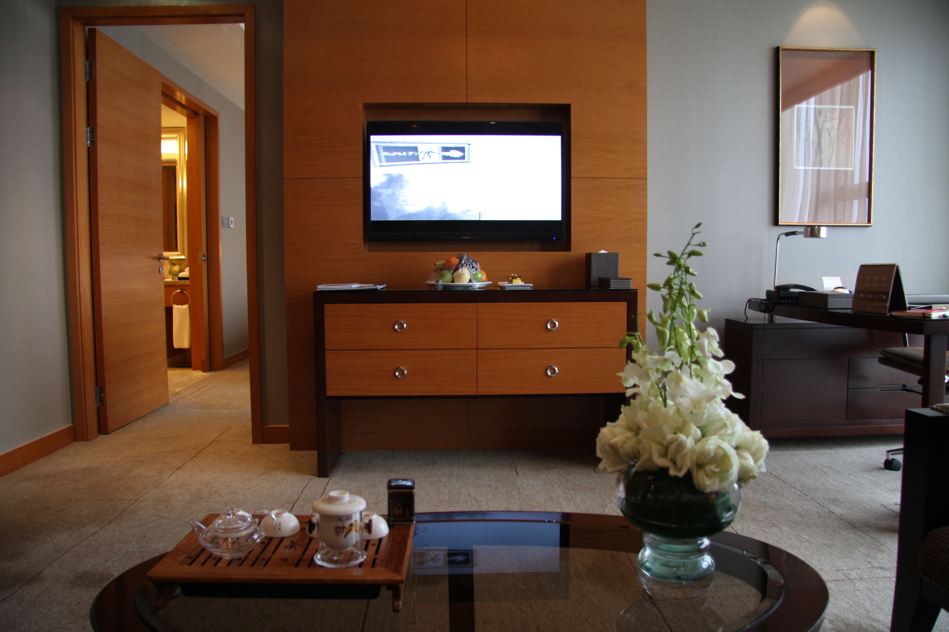 Una habitación de hotel con un armario de madera, un televisor de pantalla plana montado en la pared, un escritorio con una computadora y una mesa de centro con un jarrón de flores.