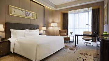 A hotel room with a large bed, two bedside tables with lamps, a desk with a chair, a window with curtains, and a decorative wall piece.