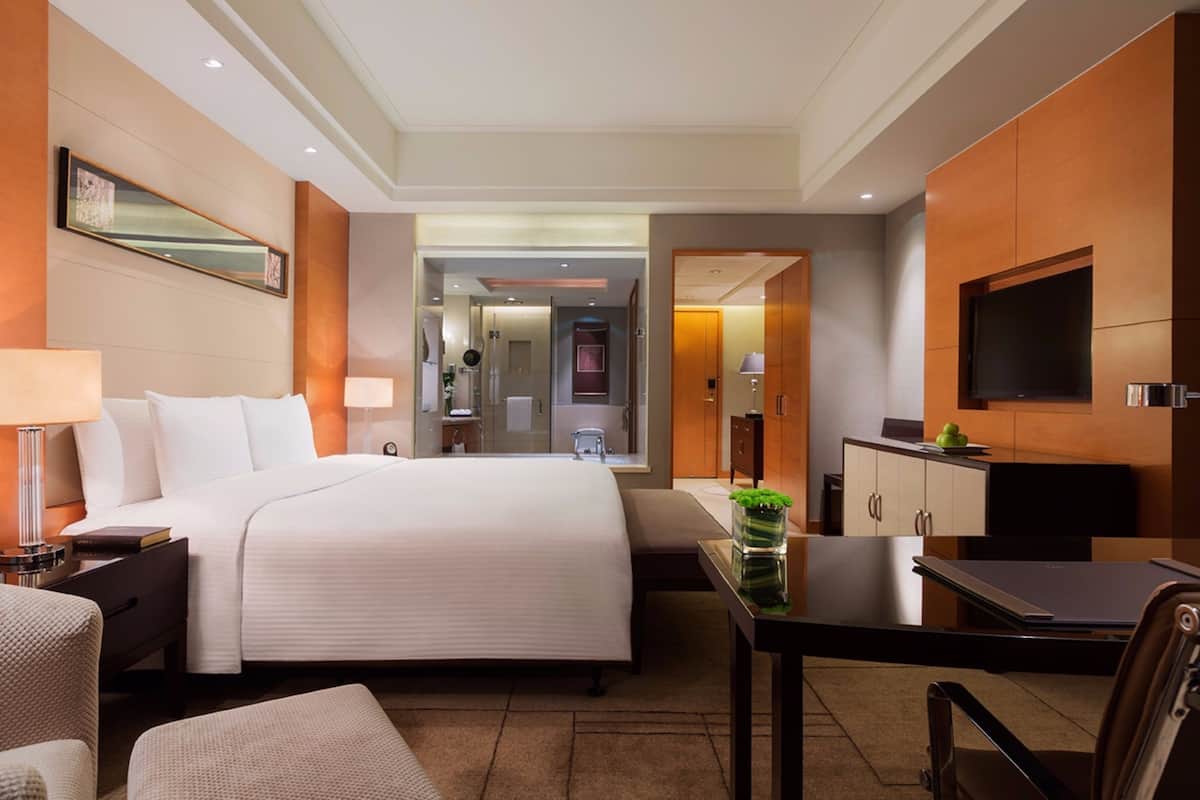A hotel room with a large bed, a desk with a laptop, a TV, a mirror, and a bathroom visible through an open door.