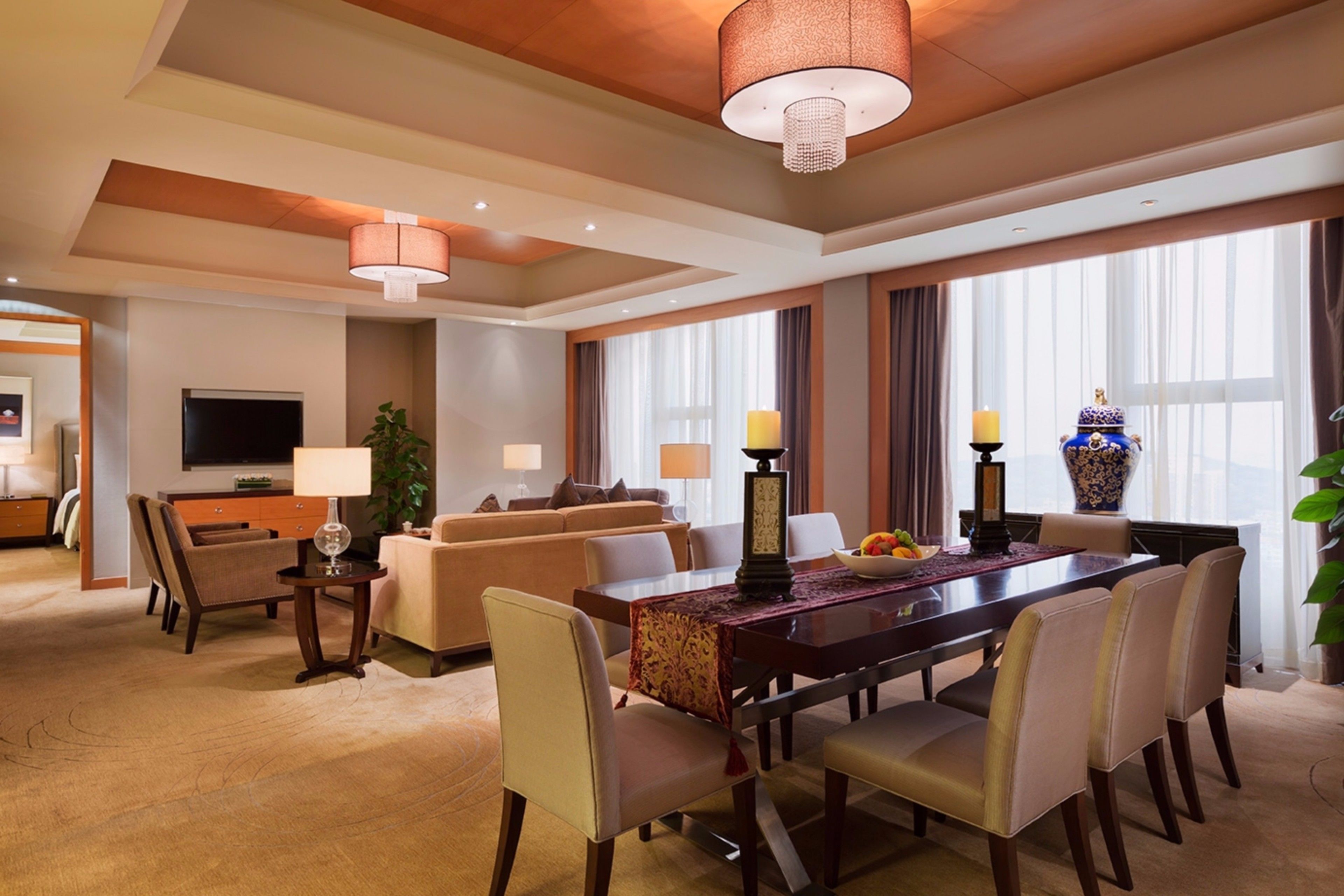 Habitación de hotel con zona de comedor, sofá, televisor y un ventanal con cortinas.