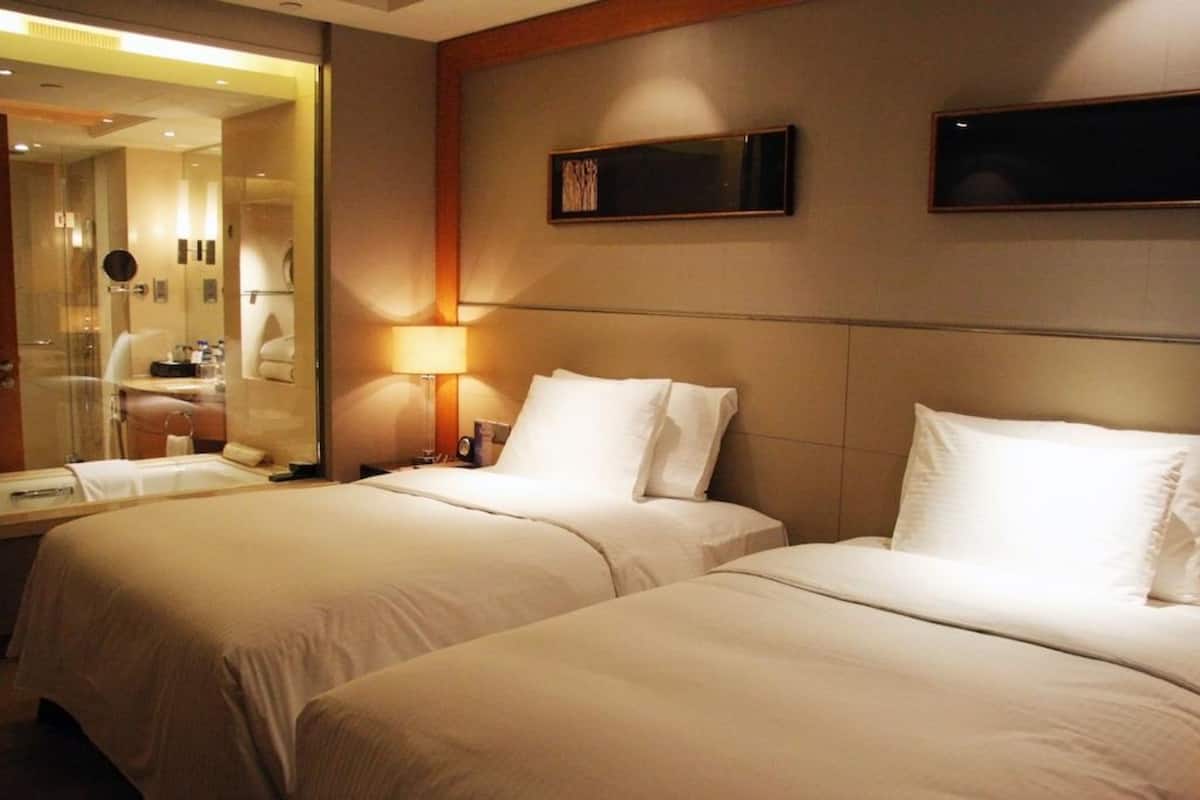 A hotel room with two beds, a large mirror, a bedside table with a lamp, and a wall-mounted TV.
