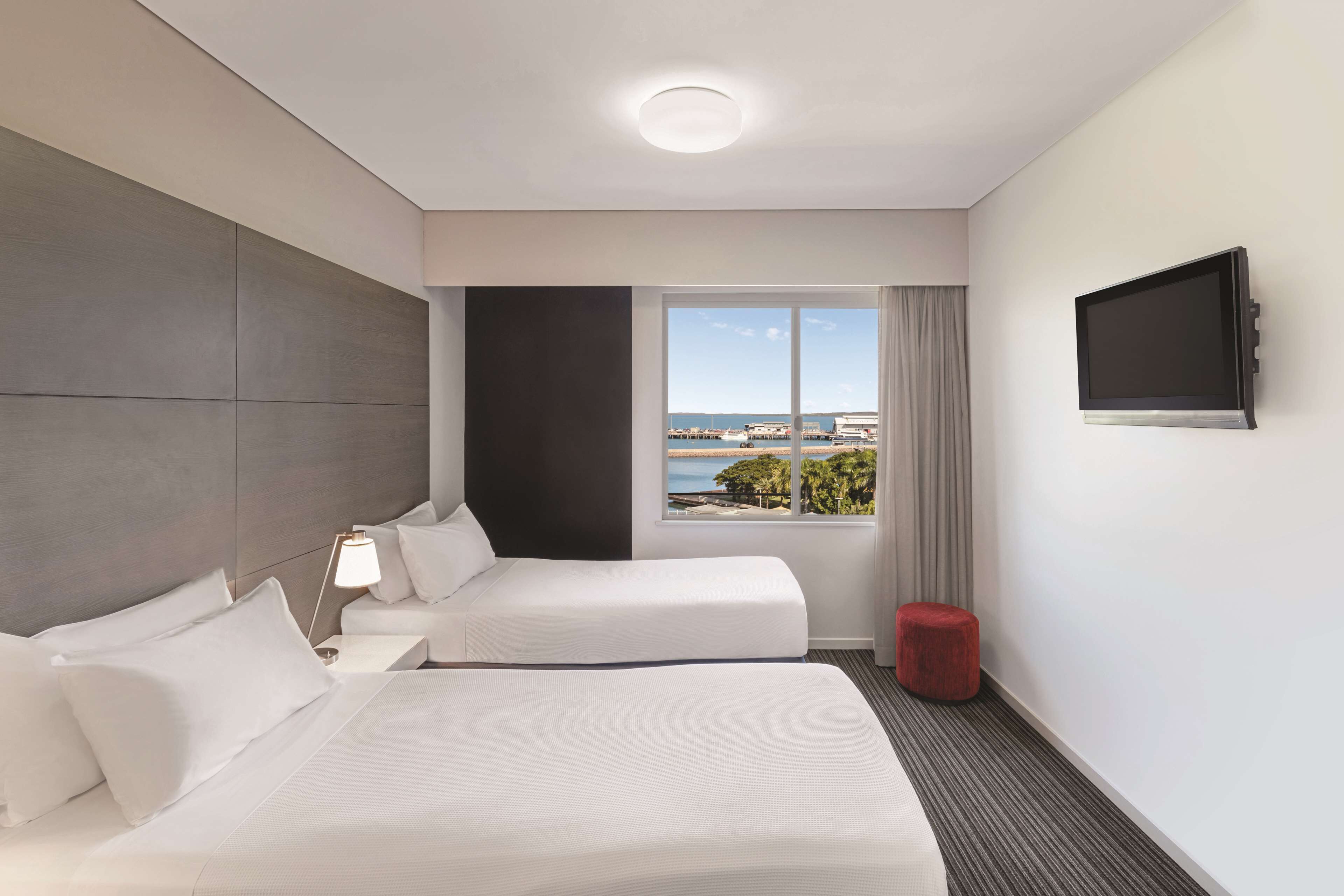 Ein Hotelzimmer mit einem Bett, einem Nachttisch mit Lampe, einem an der Wand befestigten Fernseher und einem Fenster mit Blick auf das Meer und Gebäude.