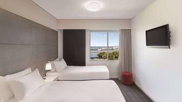Ein Hotelzimmer mit einem Bett, einem Nachttisch mit Lampe, einem an der Wand befestigten Fernseher und einem Fenster mit Blick auf das Meer und Gebäude.