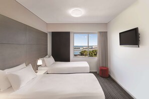 A hotel room with a bed, a bedside table with a lamp, a TV mounted on the wall, and a window with a view of the sea and buildings. - Adina Apartment Hotel Darwin Waterfront (Darwin)