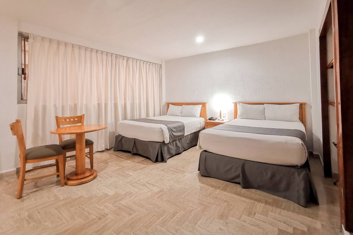 A hotel room with two beds, a small table with a chair, and a window with curtains.