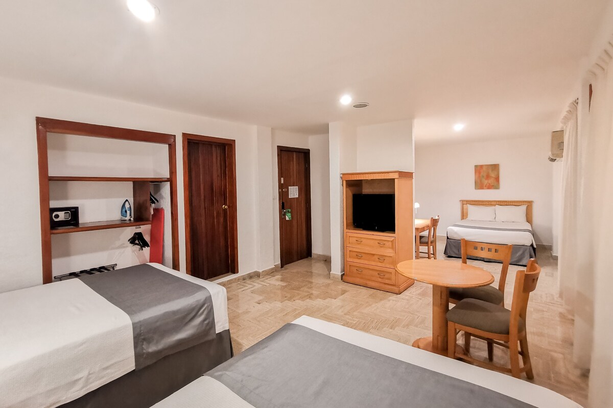 A hotel room with two beds, a wooden wardrobe, a desk with a chair, and a small table.