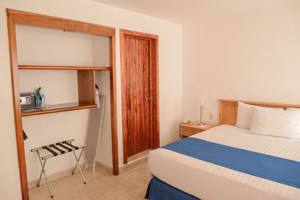 A hotel room with a bed, a wooden wardrobe, a small table with a lamp, and a drying rack.