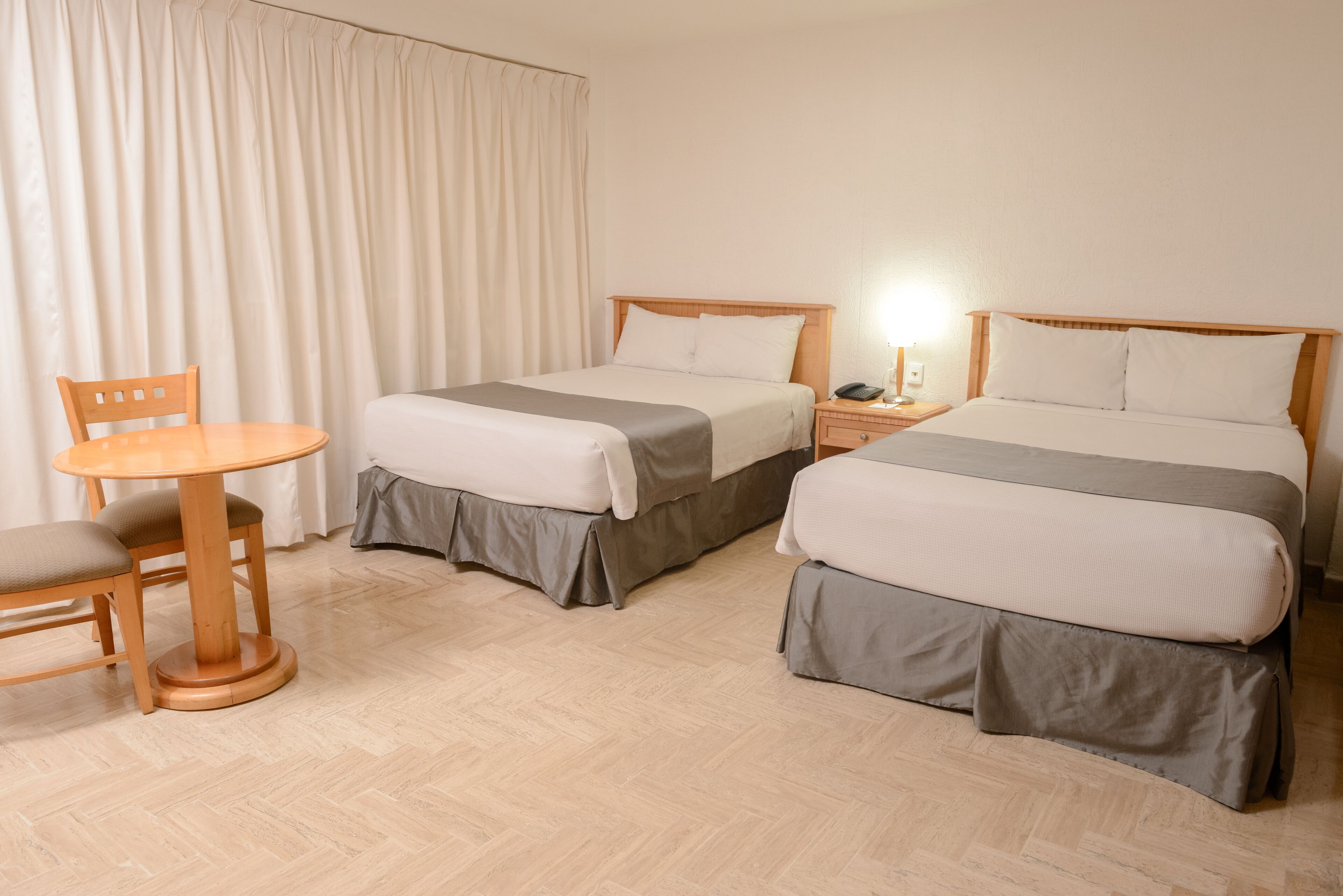A hotel room with two beds, a wooden table with a chair, a nightstand with a lamp, and a large window with curtains.