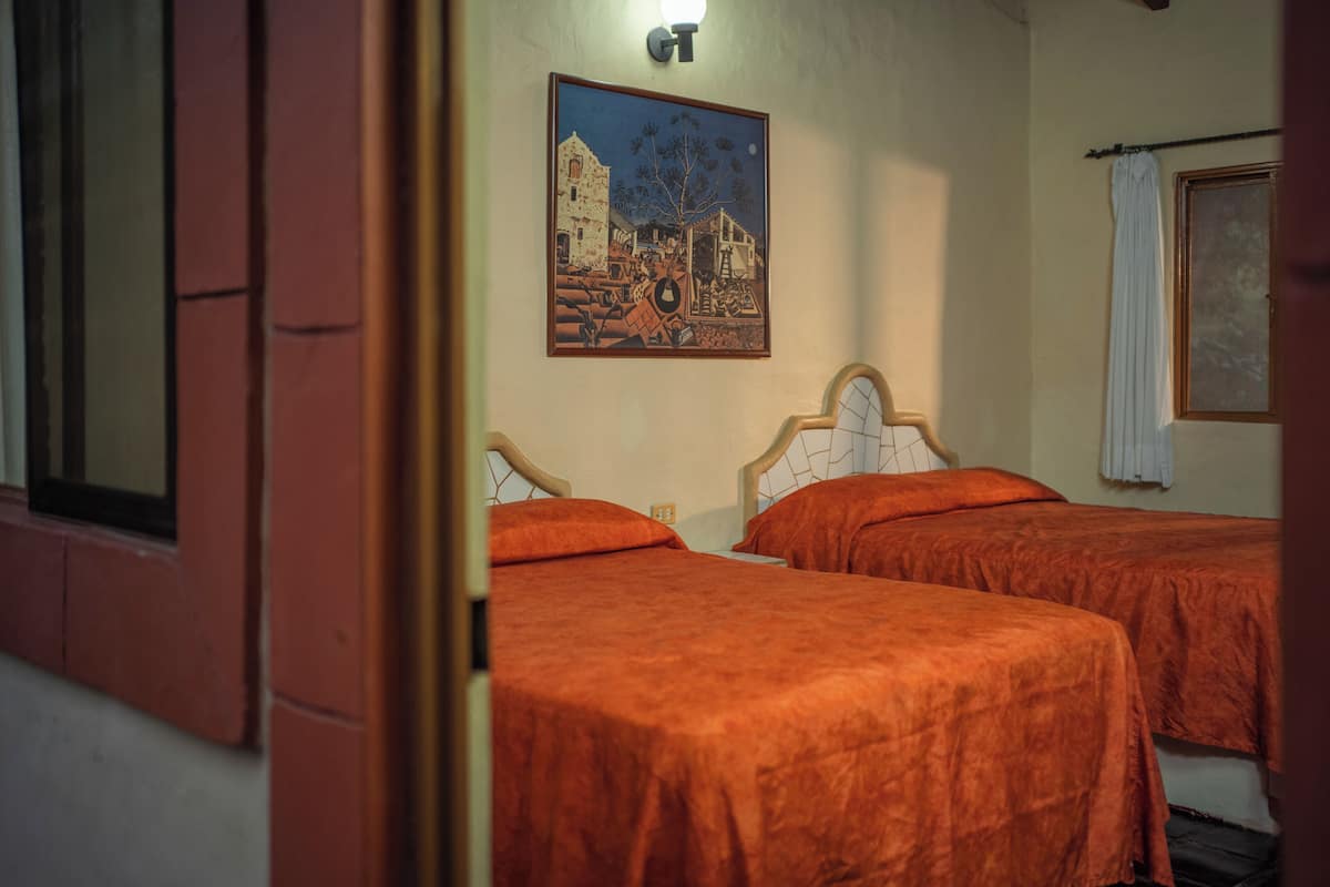 A room with two beds, a painting on the wall, and a window with a curtain.