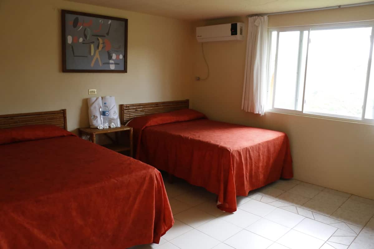 A hotel room with two beds, a painting on the wall, an air conditioning unit, and a window with curtains.